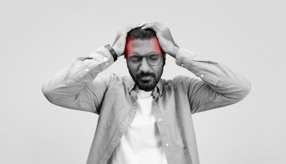 A stressed man in a casual linen shirt and glasses grasps his head in frustration or headache, with a red glow highlighting the temple area, on grey background. Headache, migraine, health problems