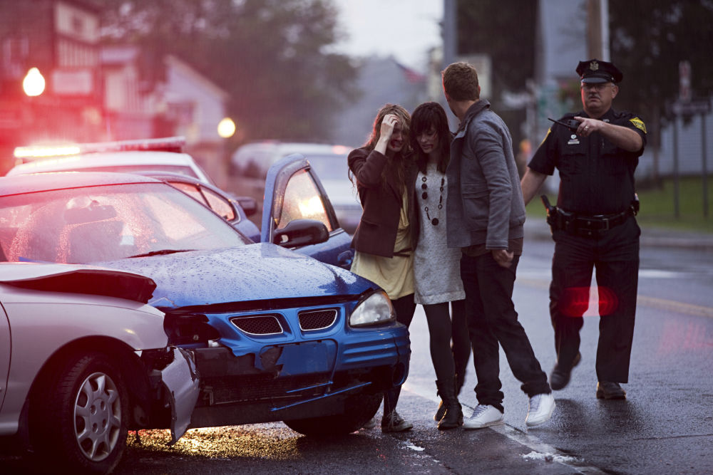 Woman comforted by man at the scene of a car accident as a police officer directs traffic.