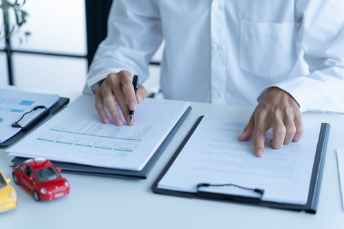 Person signing insurance forms beside toy cars and clipped documents on desk