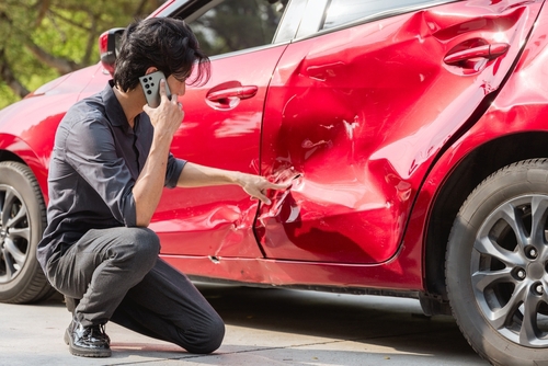 man crouching beside crash-damaged car door while talking on phone