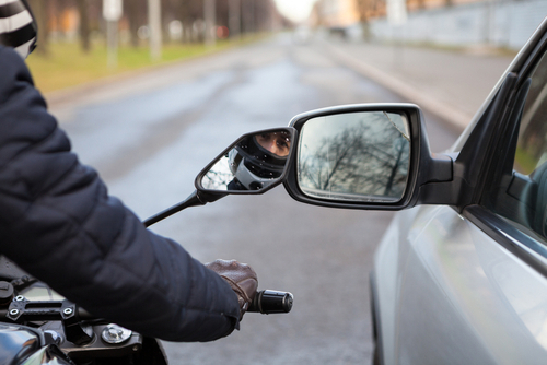 helmeted motorcycle rider touching car side mirror on road
