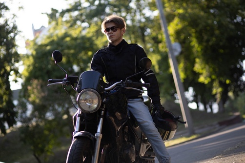 motorcyclist wearing jacket and sunglasses seated on motorcycle near tree-lined road